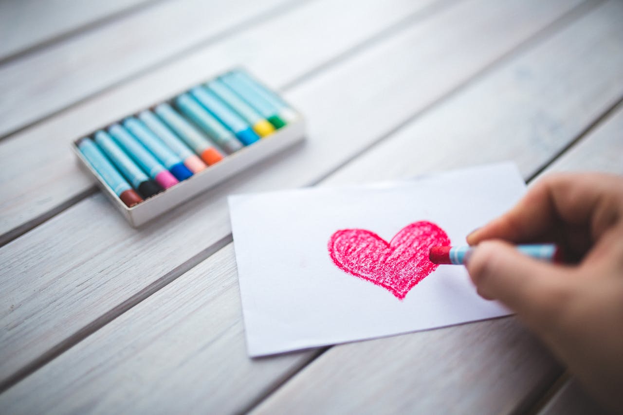A close-up of a hand using pastel crayons to draw a red heart on paper.