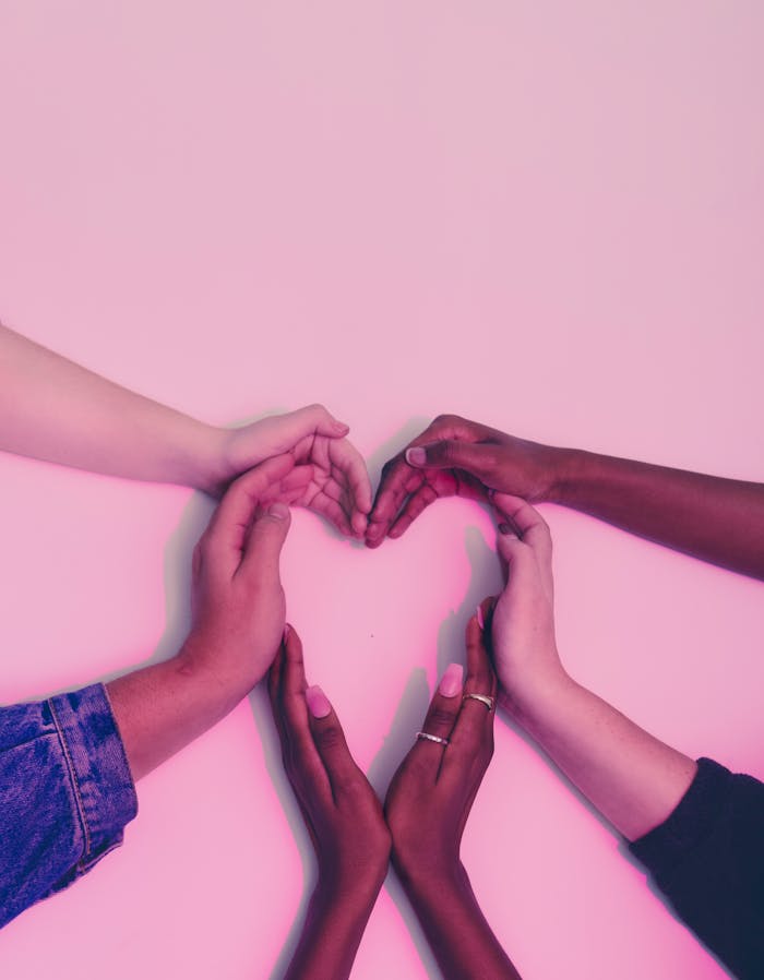 A group of diverse hands forming a heart shape against a soft pink background, symbolizing love and unity.